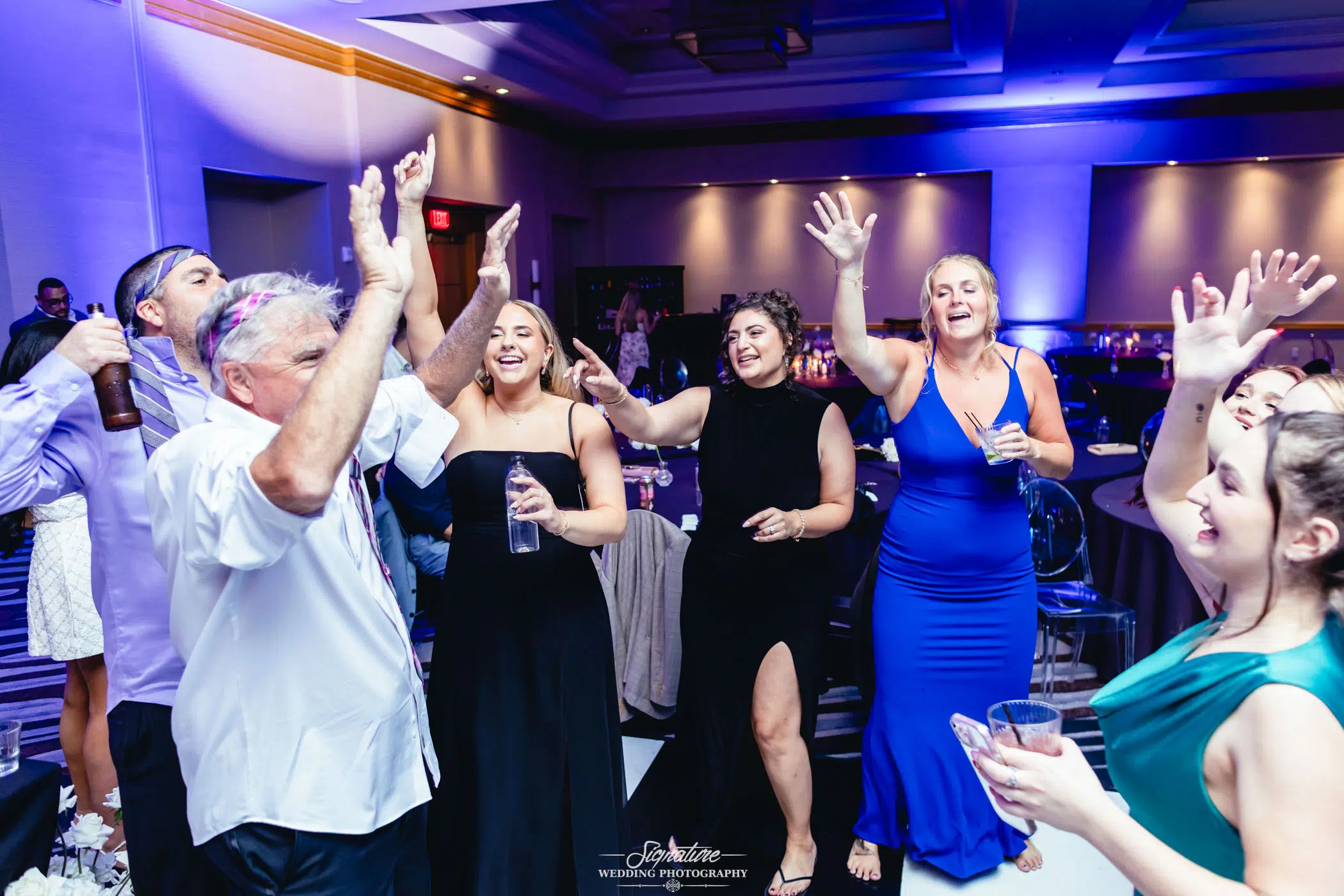 Image by Signature Wedding Photography A group of people in formal attire enthusiastically dance and raise their hands at a lively indoor party or wedding reception, drinks in hand, with tables and purple lighting in the background.