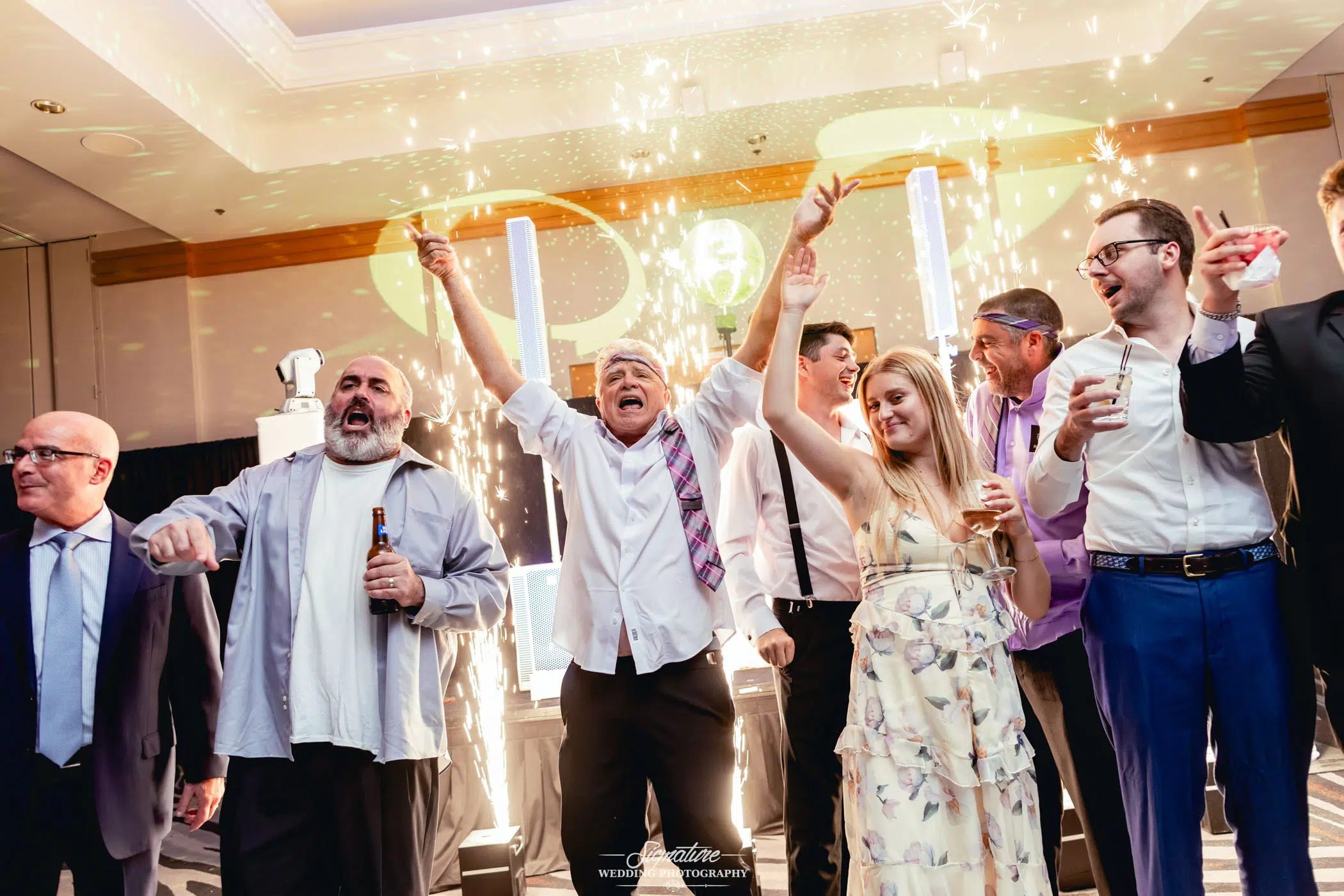 Image by Signature Wedding Photography A group of people dressed in semi-formal attire celebrate indoors, cheering and holding drinks, as bright indoor fireworks and sparklers light up the scene around them.