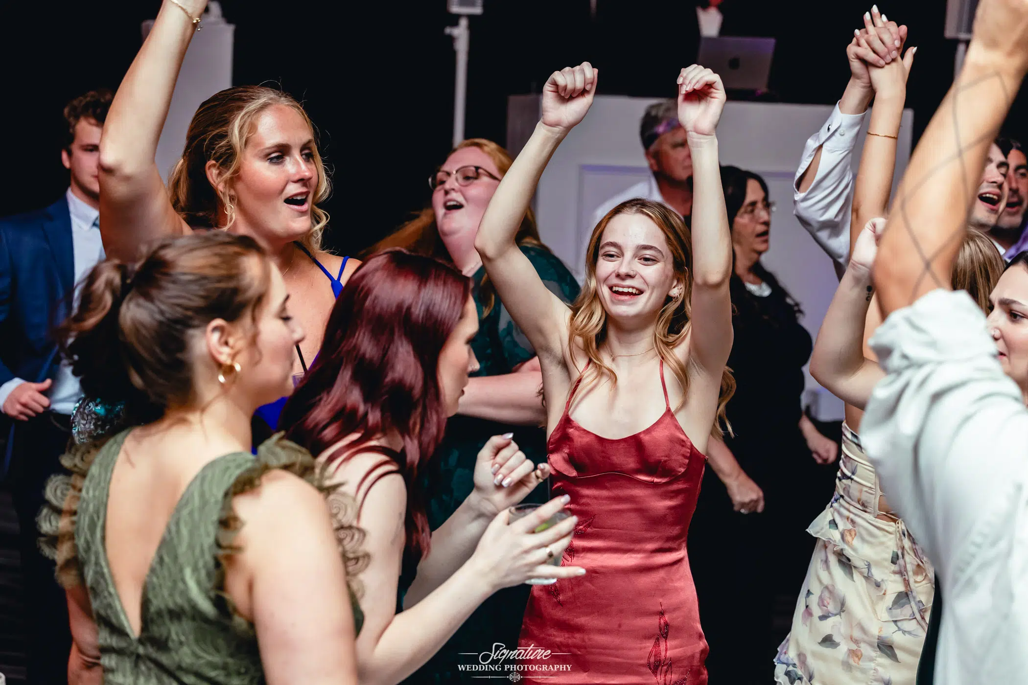 Image by Signature Wedding Photography A group of people in formal attire joyfully dancing at a celebration, with one woman in a red dress smiling and raising her arms in the air. Others around her are also cheering and enjoying the festive atmosphere.