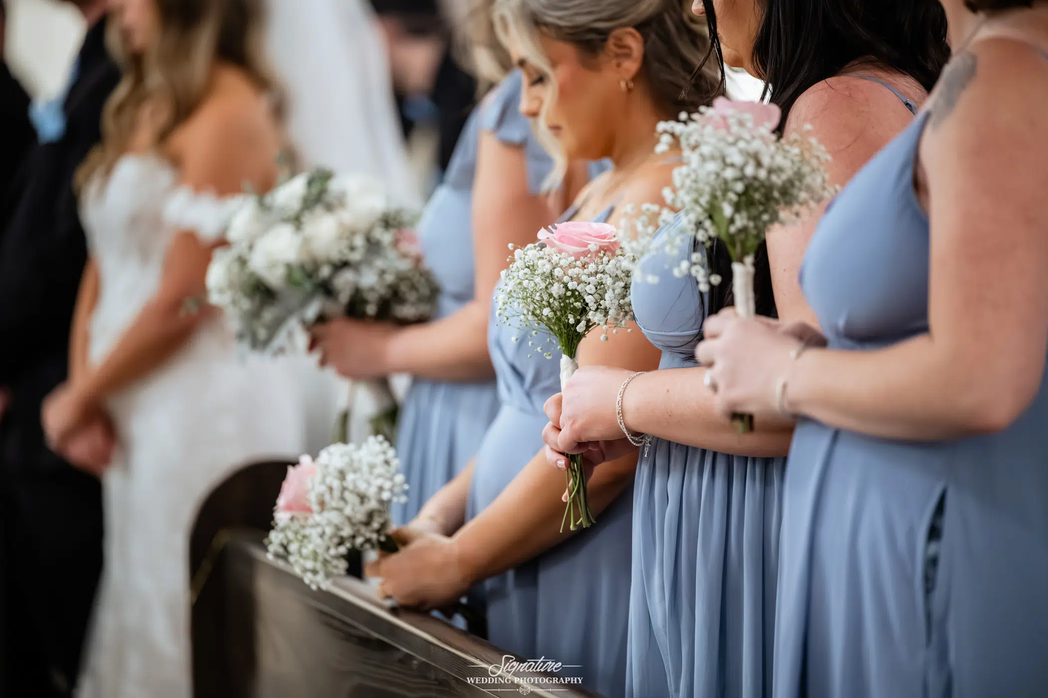 Image by Signature Wedding Photography Bridesmaids in light blue dresses hold bouquets of pink roses and baby's breath while standing in a row at a wedding ceremony. A bride in white is visible in the background.