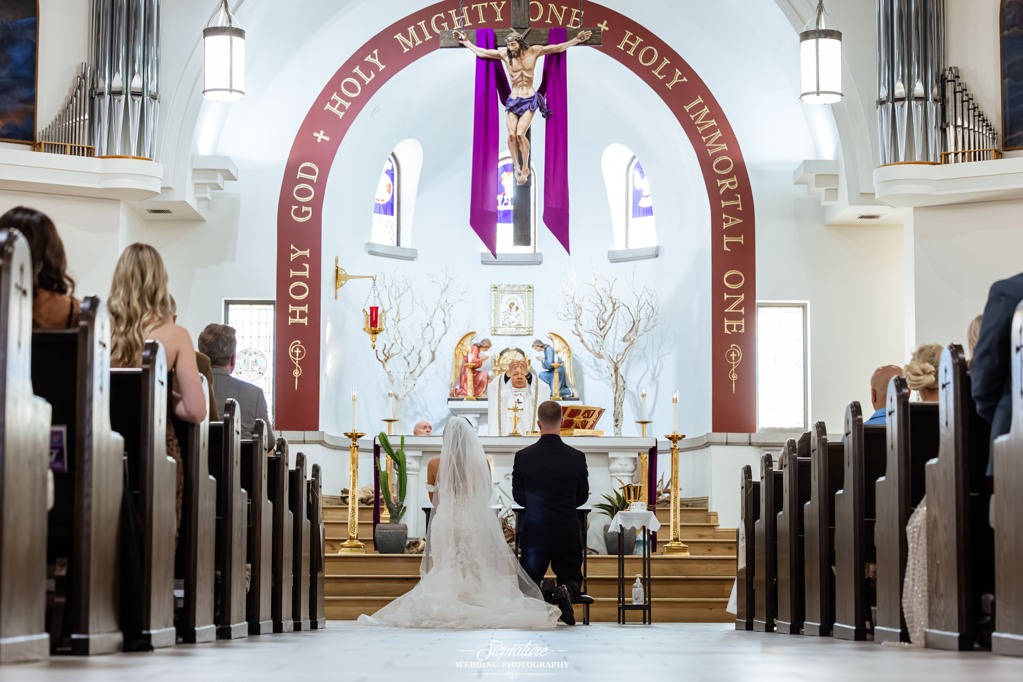 Image by Signature Wedding Photography A bride and groom kneel at the altar in a church during their wedding ceremony, surrounded by guests seated in pews. The altar is decorated with flowers, candles, and a large crucifix above.
