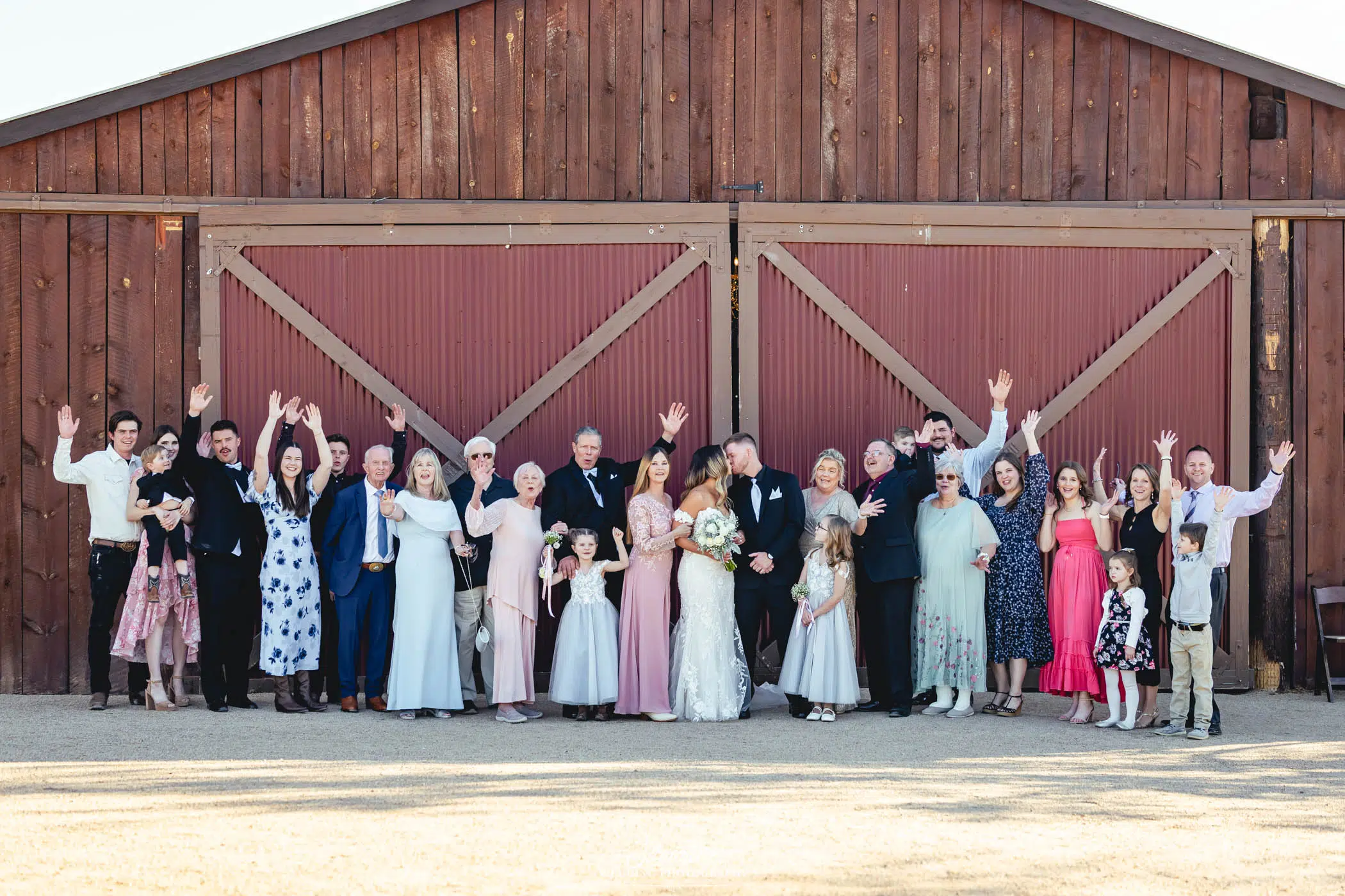 Image by Signature Wedding Photography A large wedding group poses in front of a wooden barn. The bride and groom stand in the center, surrounded by family and friends, many with arms raised, all smiling and dressed in formal attire.
