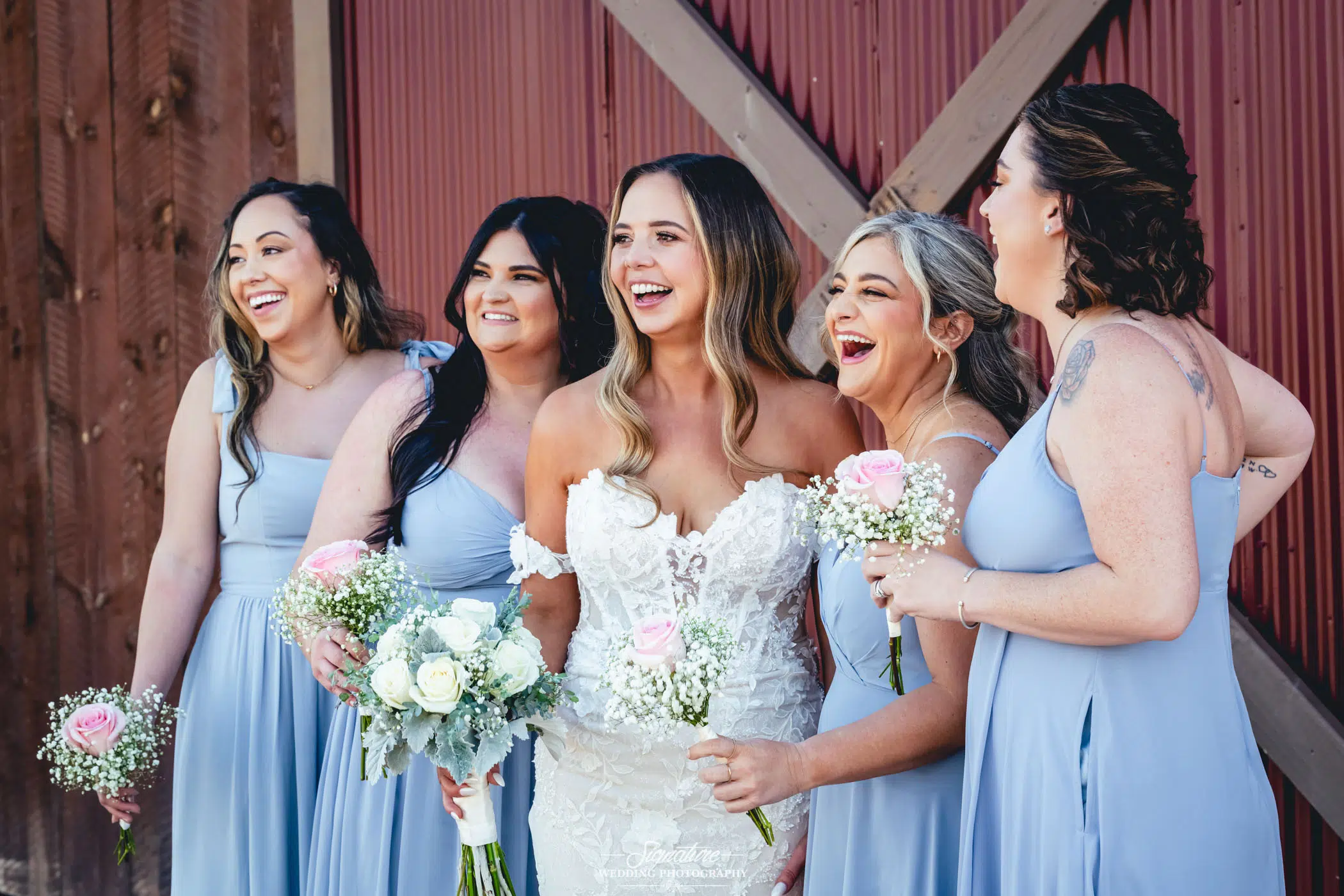 Image by Signature Wedding Photography A bride in a white gown stands with four bridesmaids in light blue dresses, all smiling and holding bouquets of white and pink flowers in front of a rustic wooden barn wall.