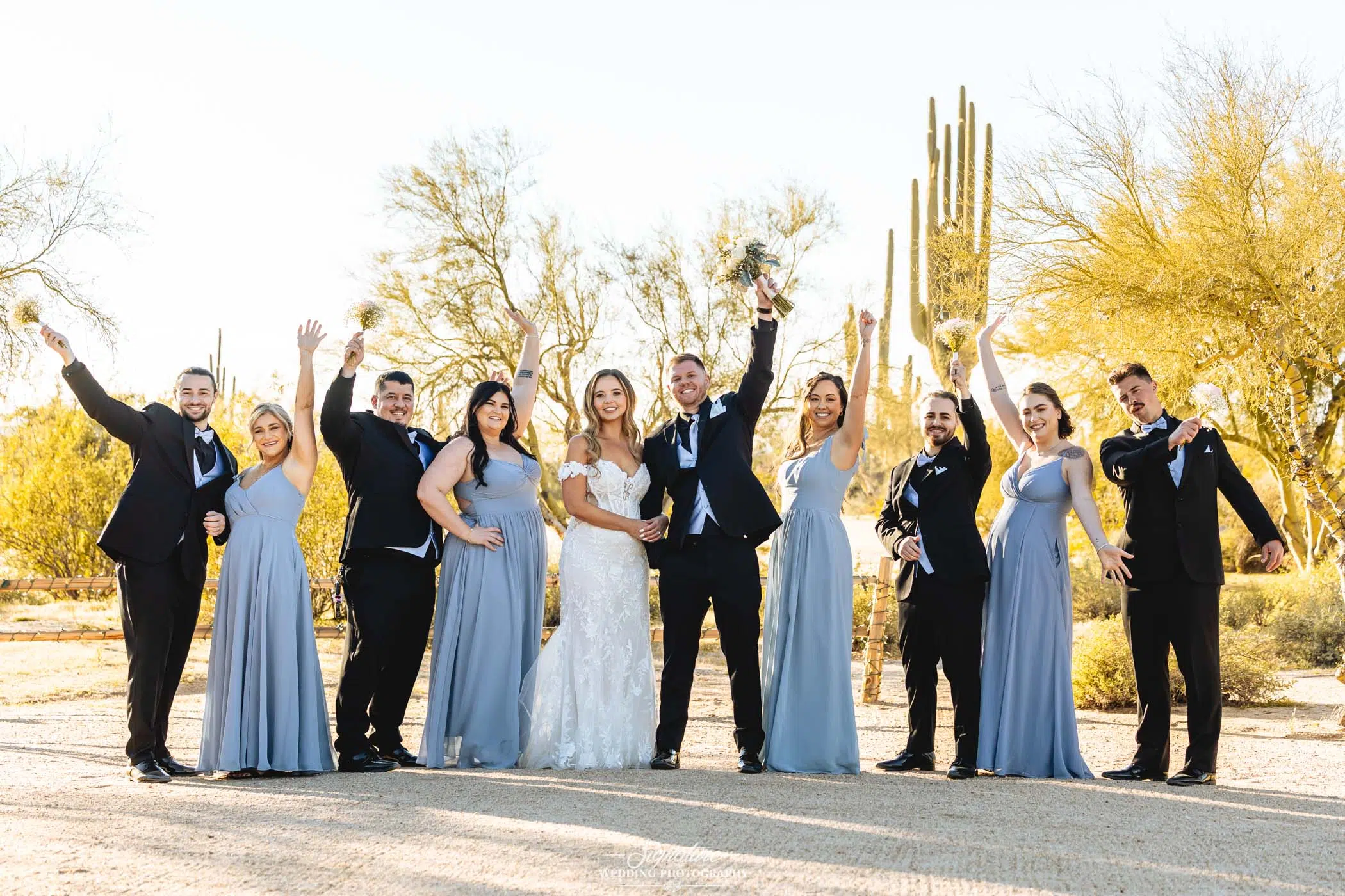Image by Signature Wedding Photography A bride and groom stand with their wedding party outdoors, surrounded by cacti and trees. The bridesmaids wear light blue dresses, the groomsmen wear black suits, and everyone is smiling and raising their arms in celebration.