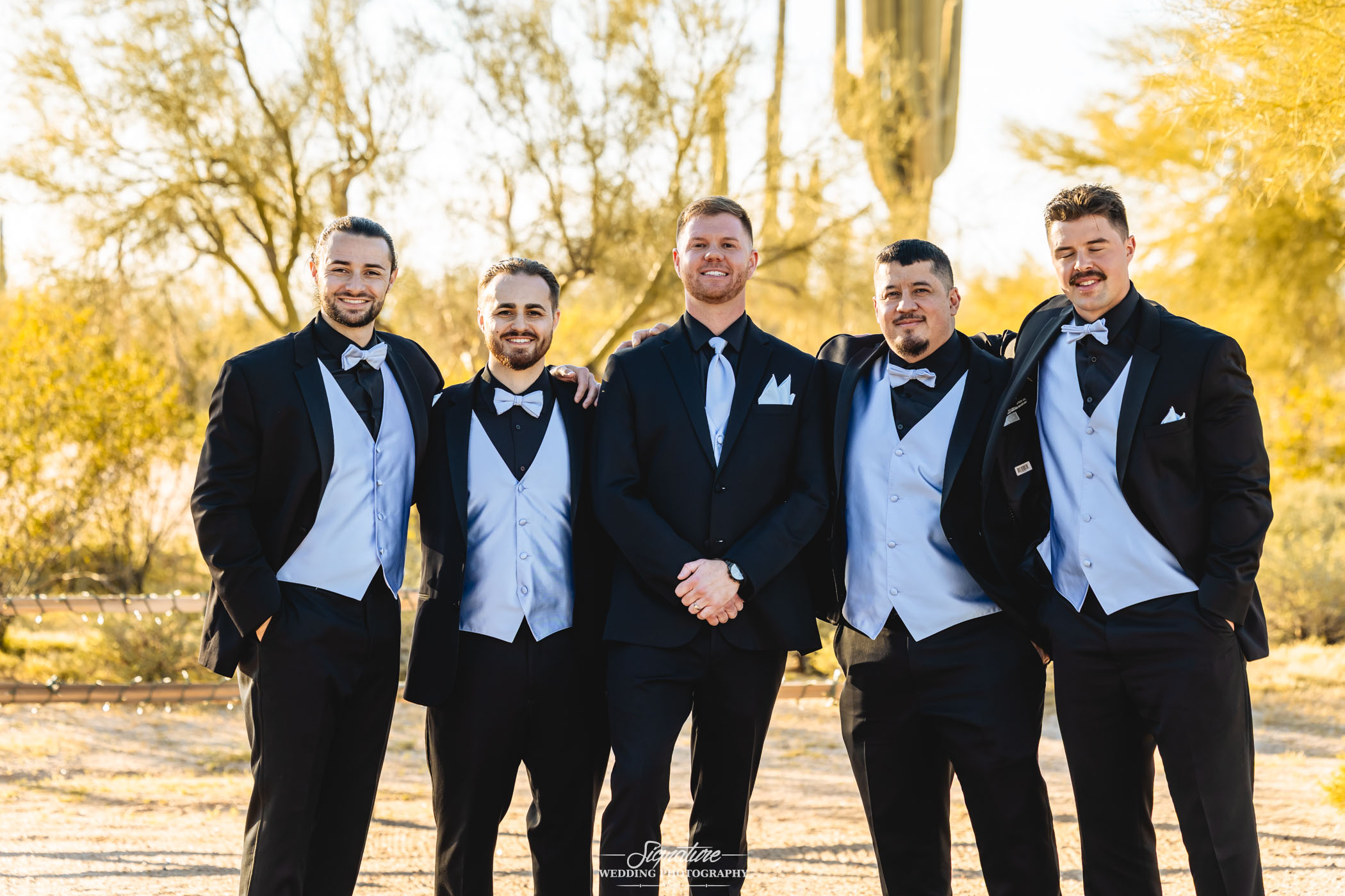 Image by Signature Wedding Photography Five men in tuxedos stand outdoors, smiling with arms around each other in a sunny, desert-like setting with trees and a cactus in the background.