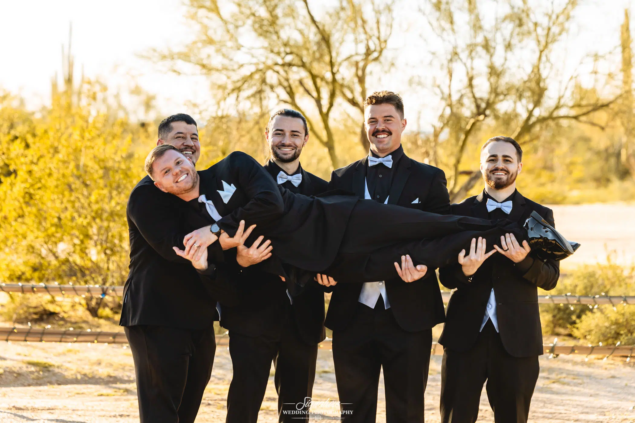 Image by Signature Wedding Photography Five men in black tuxedos pose outdoors; four stand smiling and hold up the fifth man horizontally in front of them. The background features desert vegetation and sunlight.