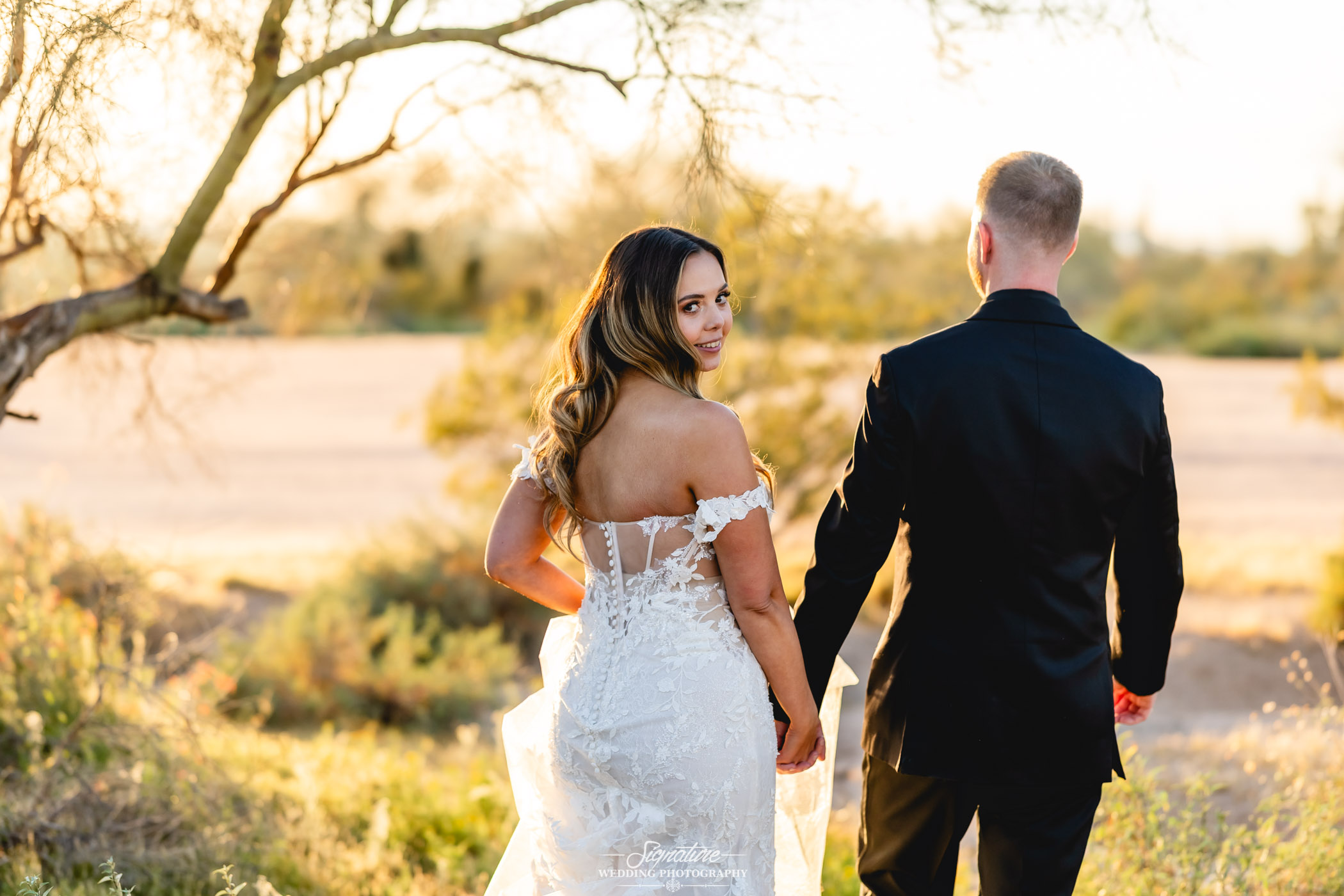 Image by Signature Wedding Photography A bride in a white lace gown looks back and smiles while holding hands with a groom in a black suit, standing outdoors in a sunlit, natural landscape.
