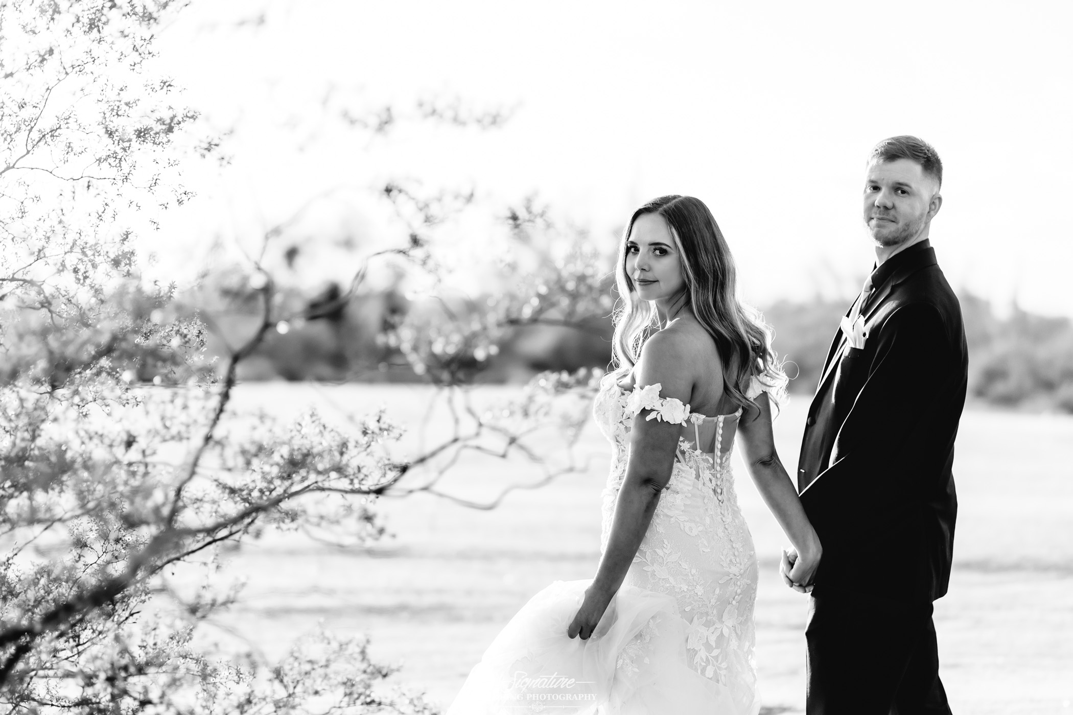 Image by Signature Wedding Photography A bride and groom hold hands and look back toward the camera while standing outdoors. The bride wears a lace wedding dress and the groom wears a suit. Trees and open field are in the background. The photo is in black and white.