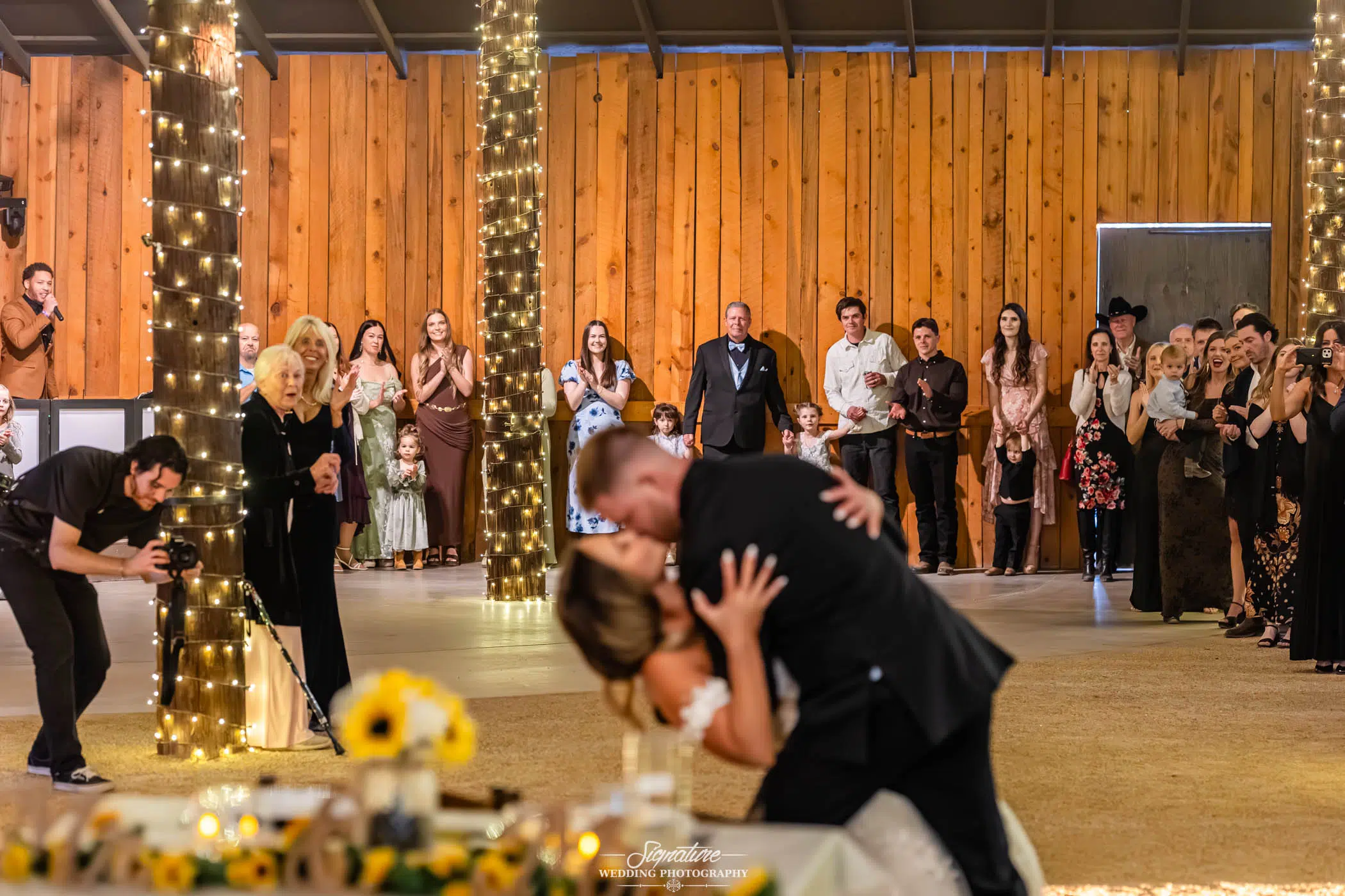 Image by Signature Wedding Photography A bride and groom share a kiss on the dance floor at their wedding reception, surrounded by guests who are clapping and smiling. The venue features wooden walls, lighted pillars, and a photographer capturing the moment.