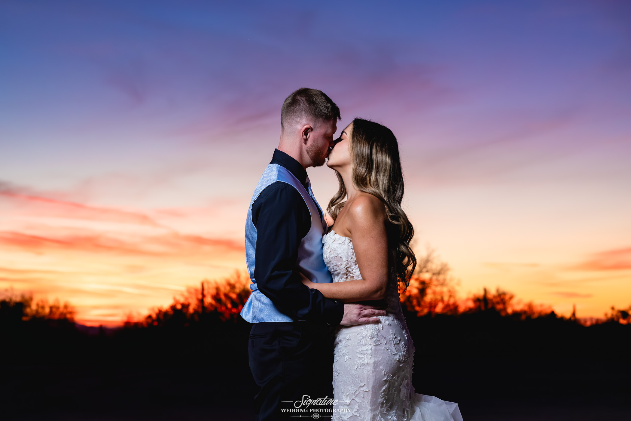 Image by Signature Wedding Photography A bride and groom stand close together at sunset, gazing into each other's eyes with their foreheads touching. The sky behind them is vibrant with orange, purple, and blue hues.