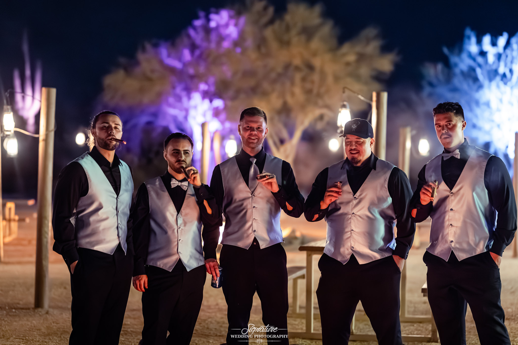 Image by Signature Wedding Photography Five men dressed in black shirts, matching silver vests, and bow ties stand outdoors at night, each holding a cigar. The background features illuminated trees with purple and yellow lighting.