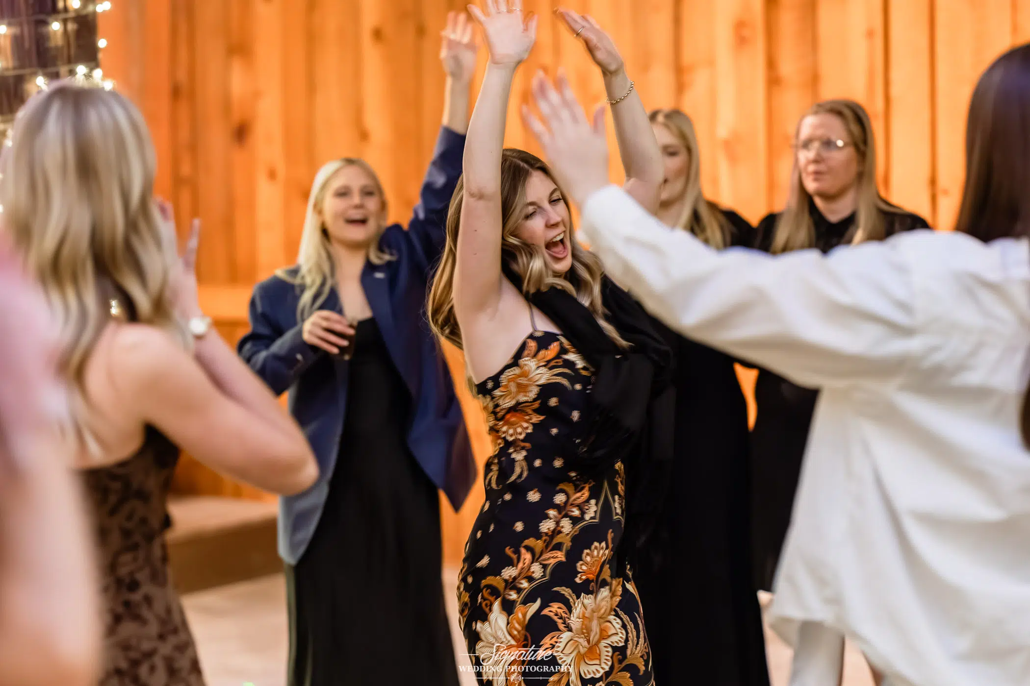 Image by Signature Wedding Photography A group of women, dressed in semi-formal attire, cheerfully dance and raise their arms in celebration inside a warmly lit wooden room. One woman in a floral dress stands out with her arms up, smiling joyfully.
